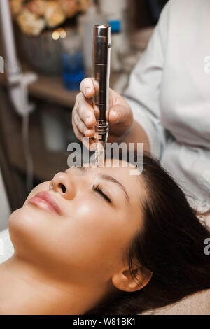 Nahaufnahme, Porträt einer schönen Frau mit dunklen Haaren mit geschlossenen Augen leanin auf einem Bett, während mit microneedling mit dermapen durch eine Kosmetikerin. Stockfoto