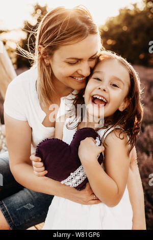 Nahaufnahme, Porträt einer schönen Mutter und Tochter umarmen und Lachen bei Kamera schaut gegen Sonnenuntergang in einem Feld mit Blumen. Stockfoto