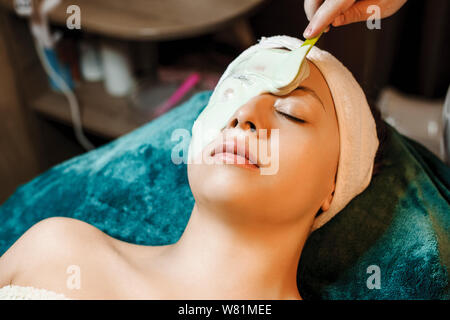 Porträt einer charmanten Frau lehnte sich auf ein Spa Bett, während auf ihrem Gesicht ist ein bio Alginat Maske durch ein Cosmetologist angewendet. Stockfoto
