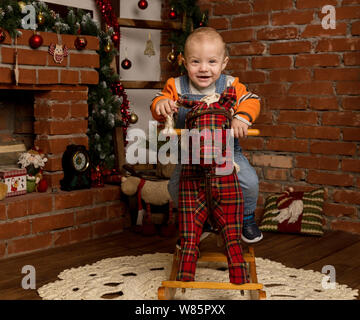 Kleines Baby auf Schaukelpferd, in Pullover und Jeans gekleidet. Weihnachten und neues Jahr Dekorationen Stockfoto