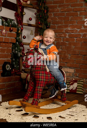 Kleines Baby auf Schaukelpferd, in Pullover und Jeans gekleidet. Weihnachten und neues Jahr Dekorationen Stockfoto