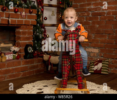 Kleines Baby auf Schaukelpferd, in Pullover und Jeans gekleidet. Weihnachten und neues Jahr Dekorationen Stockfoto