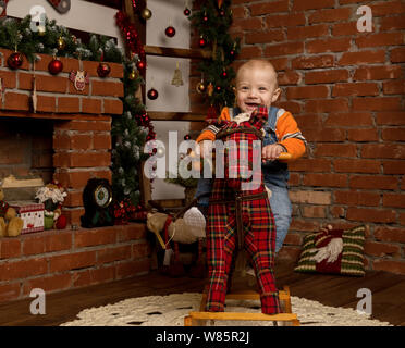 Kleines Baby auf Schaukelpferd, in Pullover und Jeans gekleidet. Weihnachten und neues Jahr Dekorationen Stockfoto