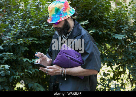 Mailand, Italien - 17. JUNI 2019: Mann mit lila Prada Tasche, schwarze Jacke und bunten Hut am Smartphone auf der Suche vor Fendi fashion show, Milan Mode Stockfoto