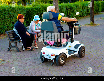 Modugno, Italien - 11. Mai 2018: Ältere Menschen auf einer Parkbank zu sozialisieren. Eine behinderte Person mit einem motorisierten Rollstuhl Stockfoto