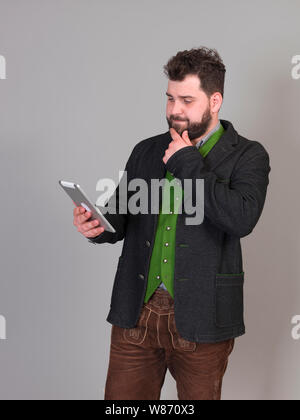 Jung und cool Österreichischer Mann mit schwarzem Haar und Bart in der traditionellen Tracht steht vor einem grauen Hintergrund und arbeitet auf einem Tablet Stockfoto