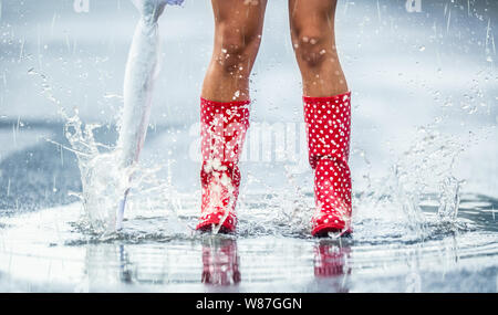 Frau Beine in gestrichelten roten Gummistiefel mit Regenschirm springen im Sommer Frühling oder Herbst Pfützen. Stockfoto