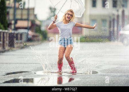 Fröhliches Mädchen springen mit weißer Schirm in punktierten roten Galoschen. Heißer Sommer Tag nach dem regen Frau springen und Plantschen in der Pfütze Stockfoto