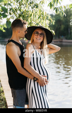 Foto von Schwangeren blonde Frau in Hut und Männer in rosa Brille stehen am Ufer des Flusses Stockfoto