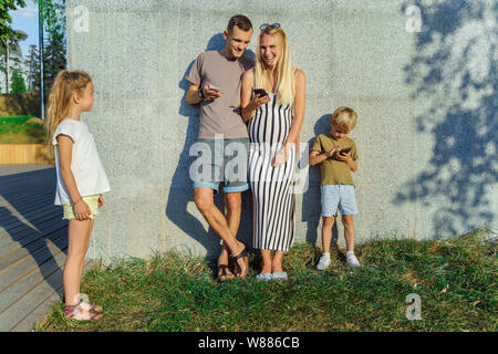 Foto von Frau, Mann und Sohn mit Telefonen in den Händen und Töchter neben Beton Wand außerhalb Stockfoto