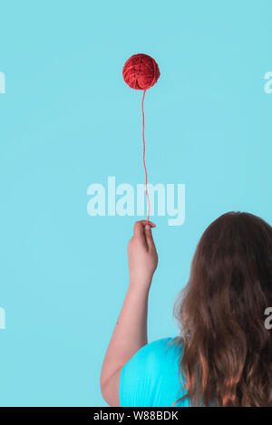 Seltsame Kindheit Konzept mit einer Frau mit einem schwebenden roten Faden Kugel wie ein Ballon. Sonderbar stricken Handarbeiten Konzept. Minimale Kindheit frame Stockfoto