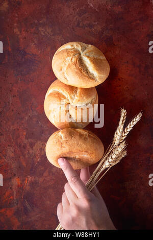 Pyramide der rostigen runde Brötchen (Kaiser oder Wien Rollen), Ausgleich auf jedem anderen mit der Hand hält Weizenähren auf dunklen Rot strukturierten Hintergrund Stockfoto