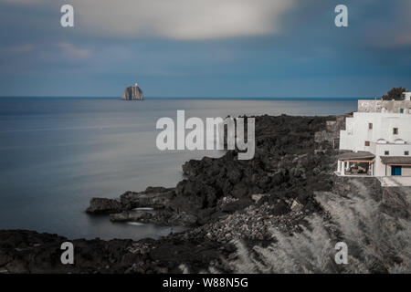 Ein Leuchtturm auf einer kleinen Insel, von einer Klippe mit schwarzen Felsen und ein weißes Gebäude gesehen. Sizilien, Italien. Stockfoto