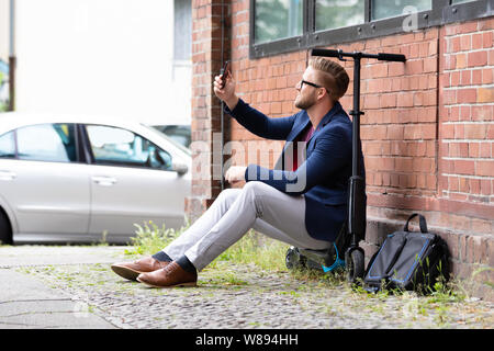 Lächeln, Geschäftsmann, Sitzen auf elektrische Motorroller in der Nähe von Mauer unter Selfie mit Handy Stockfoto