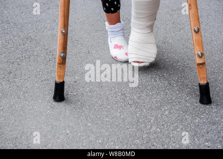 Junge Mädchen in der orthopädischen cast auf Krücken zu Fuß auf der Straße in der Nähe der Straße. Kind mit einem gebrochenen Bein auf Krücken, knöchelverletzung. Knochenbruch und Stockfoto