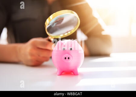 Nahaufnahme einer Geschäftsfrau, Prüfung der rosa Sparschwein durch Lupe auf weißem Schreibtisch Stockfoto