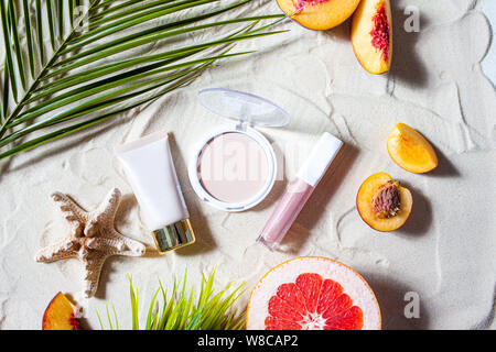 Bezaubernden Strand. Dekorative Kosmetik in Pastellfarben, Pulver, Basis, Lip Gloss mit hellen Früchten, Seesterne, und Palm Blätter liegen auf dem weißen Sand. Stockfoto