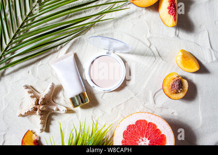 Bezaubernden Strand. Dekorative Kosmetik in Pastellfarben, Pulver, Basis, Lip Gloss mit hellen Früchten, Seesterne, und Palm Blätter liegen auf dem weißen Sand. Stockfoto