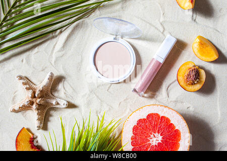 Bezaubernden Strand. Dekorative Kosmetik in Pastellfarben, Pulver, Basis, Lip Gloss mit hellen Früchten, Seesterne, und Palm Blätter liegen auf dem weißen Sand. Stockfoto