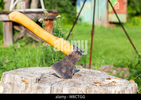 Nahaufnahme einer großen Beil mit Holzgriff in einem hölzernen Stumpf nach dem Zerkleinern von Brennholz auf grünem Hintergrund an einem warmen Sommertag klemmt Stockfoto