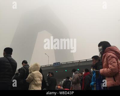 Fußgänger, von denen die meisten Gesicht-masken tragen, warten Sie eine Straße vor der neue Hauptsitz der CCTV-vage in schweren Smog in Peking, China gesehen, Stockfoto