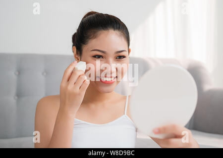 Schöne asiatische Frau mit kosmetischen Schwamm auf Gesicht und Spiegel zu Hause aussehen Stockfoto