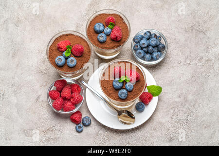 Classic Tiramisu Dessert mit Blaubeeren und Himbeeren in ein Glas und Schüsseln mit Beeren auf konkreten Hintergrund Stockfoto