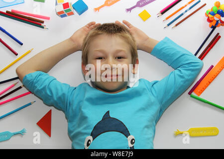 Der Junge liegt auf einem weißen Hintergrund, Schulsachen sind in der näheren Umgebung verstreut. Der Junge ist immer bereit, um in die Schule zu gehen. Lineale, Kugelschreiber, Bleistifte. Das Kind r Stockfoto