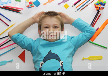 Der Junge liegt auf einem weißen Hintergrund, Schulsachen sind in der näheren Umgebung verstreut. Der Junge ist immer bereit, um in die Schule zu gehen. Lineale, Kugelschreiber, Bleistifte. Das Kind r Stockfoto