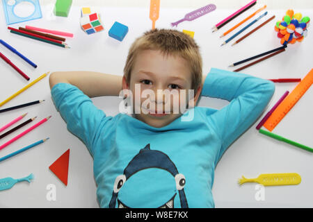 Der Junge liegt auf einem weißen Hintergrund, Schulsachen sind in der näheren Umgebung verstreut. Der Junge ist immer bereit, um in die Schule zu gehen. Lineale, Kugelschreiber, Bleistifte. Das Kind r Stockfoto