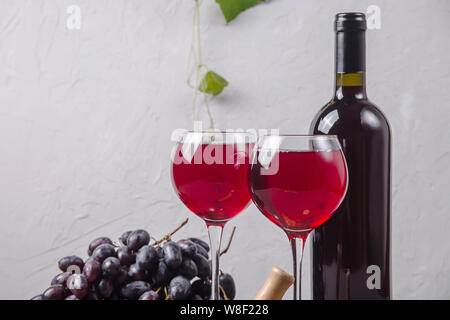 Zwei Gläser Rotwein neben einer Rolle, eine Weintraube und ein Korkenzieher auf einem Hintergrund von grünen Zweigen der Trauben auf grauem Hintergrund Stockfoto