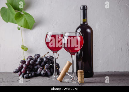 Zwei Gläser Rotwein neben einer Rolle, eine Weintraube und ein Korkenzieher auf einem Hintergrund von grünen Zweigen der Trauben auf grauem Hintergrund Stockfoto