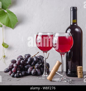 Zwei Gläser Rotwein neben einer Rolle, eine Weintraube und ein Korkenzieher auf einem Hintergrund von grünen Zweigen der Trauben auf grauem Hintergrund Stockfoto