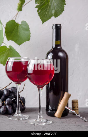 Zwei Gläser Rotwein neben einer Rolle, eine Weintraube und ein Korkenzieher auf einem Hintergrund von grünen Zweigen der Trauben auf grauem Hintergrund Stockfoto