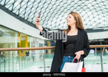 Schöne braunhaarige Mädchen mit Einkaufstaschen nimmt selfie in Mall Stockfoto