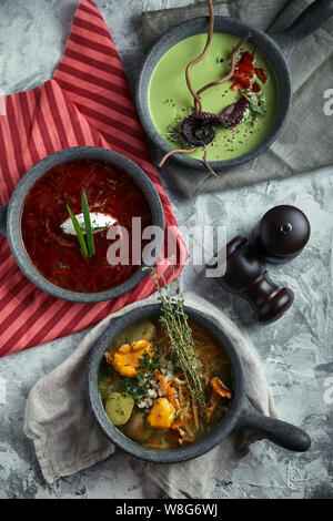 Grauen Platten mit verschiedenen Suppen auf grauem Hintergrund. Mit Spargel Suppe mit Octopus Platte, eine Platte der traditionellen Borschtsch mit saurer Sahne, eine Platte Stockfoto