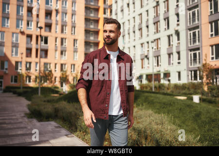 Sonnigen Tag. Hübscher junger bärtiger Mann in Freizeitkleidung zu Fuß durch die Stadt und Lächeln. Wochenende. Männer Schönheit. Lebensstil Stockfoto