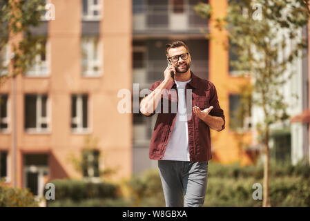 Was für ein toller Tag glücklicher bärtiger Mann in lässiger Kleidung und Brillen, der per Telefon sprach und lächelte, während er durch die Stadtstraße ging. Lifestyle. Wochenende. Kommunikation Stockfoto