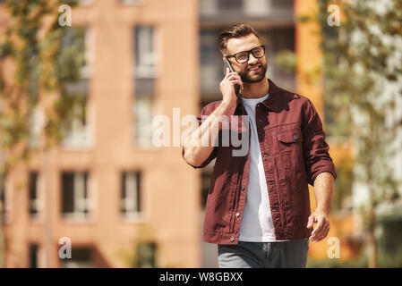 Gute Gespräche. Glücklicher bärtiger Mann in lässiger Kleidung und Brillen, der per Telefon telefoniert und lächelt, während er durch die Stadtstraße läuft. Lifestyle. Wochenende. Kommunikation Stockfoto