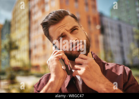 Hm... Portrait von nachdenklich bärtiger Mann sprechen über das Telefon und seine Stoppeln berühren beim Stehen auf der Straße. Schlechte Nachrichten. Digital. Kommunikation Stockfoto