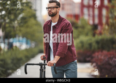 Gute Fahrt. Stattlicher Mann mit Stoppeln in legere Kleidung und Brillen sein Fahrrad ziehen beim Stehen im Freien. Wochenende Tätigkeit. Lebensstil Stockfoto