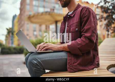 Kostenloses wi-Fi. Cropped Foto von jungen schönen Mann mit Stoppeln in Freizeitkleidung arbeiten auf Laptop, während auf der Bank im Freien sitzen. Bloggen. Stadtleben Stockfoto