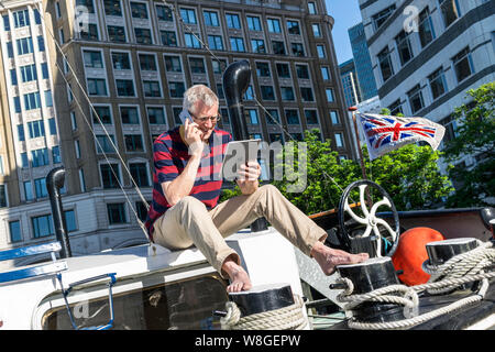 Reife erfolgreicher Unternehmer Unternehmer entspannt im Freien arbeiten auf seinem Hausboot Office über sein Smartphone iPhone an seinem iPad suchen sitzen, Büros hinter Canary Wharf London. Flagge im Wind fliegen mit British business Brexit Optimismus Konzept Stockfoto