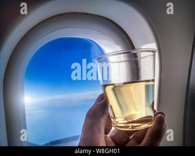 Fliegen Alkohol Getränke nervös Flyer in-flight Trinken Konzept, mit Hand Weißwein in Airline Service Kunststoff Glas, mit Kabine und blauer Himmel mit Sonne brechen in b/g Stockfoto