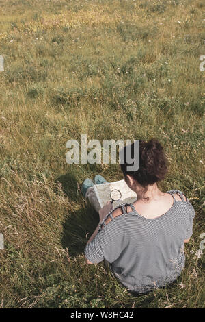 Ein Mädchen Tourist in einem gestreiften Pullover sitzt in einem Feld unter dem Gras und schaut auf eine Karte durch eine Lupe. Die Sonne scheint. Stockfoto