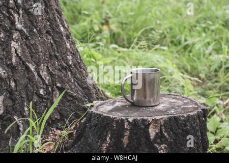 Ein Bügeleisen touristische Becher steht auf einem Baumstumpf im Wald neben einem dicken Baum vor dem Hintergrund unscharf Gras. Selektiver Fokus auf den Becher. Stockfoto