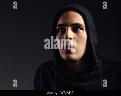 Frighting Mann in Schwarz Haube auf dem Kopf mit großen Augen schauen auf dunklem Hintergrund schockiert. Gefährliche Situation in den Schatten. Getönten portrait Stockfoto