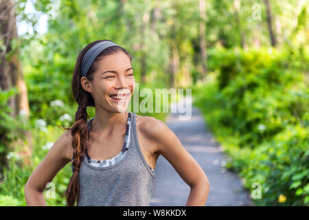 Sport asiatische Mädchen lächelnd glücklich mit ihr laufen. Runner Frau zufrieden das gute Gefühl nach dem Joggen im City Park auf Waldweg. Sportlerin tragen Fitness grau Tank Top activewear und Kopfbügel Stockfoto