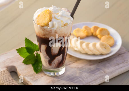 Lecker Milchshake trinken in Glas Becher mit Kunststoffrohr und Scheibe Banane auf Schneidebrett aus Holz Tisch Stockfoto