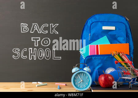 Zurück zu Schule Konzept, Bücher, Lehrbücher, Rucksack, Wecker, Apple und Schreibwaren Bürobedarf auf Klassenzimmer Schreibtisch mit schiefertafel Hintergrund. Stockfoto